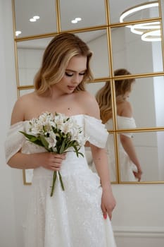 Portrait of a blonde bride with a bouquet of white flowers against a mirror. The blonde poses in a flowing wedding dress, holding white lilies. In a light interior, against a large mirror
