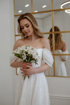 Portrait of a blonde bride with a bouquet of white flowers at the window. Blonde in a flowing wedding dress. In a light interior against the background of a window. With long hair