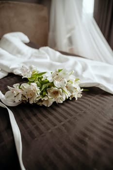 Wedding details. Bridal accessories. Flowers in the foreground. An elegant white wedding dress and a bouquet of lilies lie on a dark bed, a luxurious veil is in the background