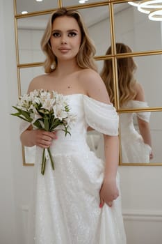 Portrait of a blonde bride with a bouquet of white flowers against the background of a mirror. Blonde in a flowing wedding dress. In a light interior, smiling happily