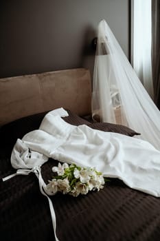 Wedding details. accessories for the bride. On the dark bed lies an elegant white wedding dress and a bouquet of lilies, in the background is a luxurious veil,