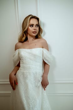 A stunning bride poses elegantly in a flowing wedding dress on a white background in a minimalist interior. She radiates grace and beauty. High-quality photo