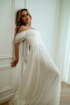 A stunning bride poses elegantly in a flowing wedding dress on a white background in a minimalist interior. She radiates grace and beauty. Blonde with long hair poses elegantly. High quality photo
