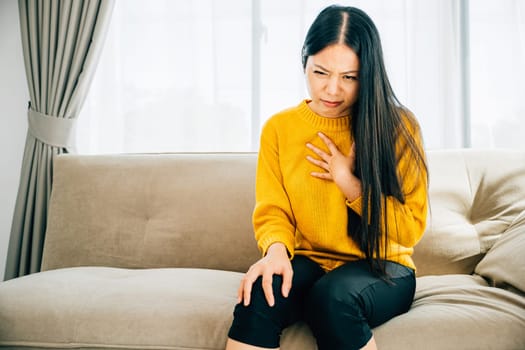Highlighting a heart attack, Asian woman on a sofa clutches her left chest in pain needing immediate medical attention. Illustrating heartache discomfort and the urgency for emergency aid.