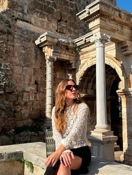 A woman sits elegantly on a stone ledge near ancient ruins, basking in the sunlight under a clear blue sky.