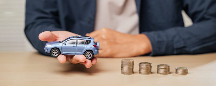 Hand holding a blue toy car model next to ascending coin stacks. Depicts auto credit, car loans, cash installment payments, and financial planning for managing car purchase costs.