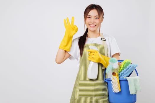 Cleaning Asian woman in striped apron holding cleaning bucket supplies, wearing gloves, shows OK finger gesture with confidence in her cleaning profession. isolated studio white background
