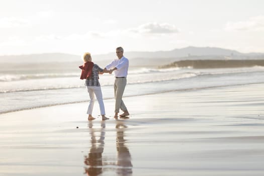 Carefree senior couple holding hands and dancing together on the beach at sunset, enjoying retirement