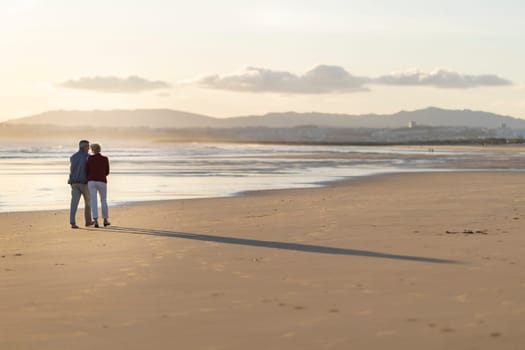 Elderly couple enjoying a romantic walk on the beach at sunset, creating a heartwarming scene of love and companionship
