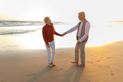 Elderly couple enjoying a romantic walk on the beach at sunset, holding hands and smiling