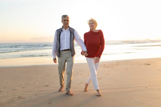 Elderly couple holding hands and walking barefoot on the beach at sunset, enjoying retirement