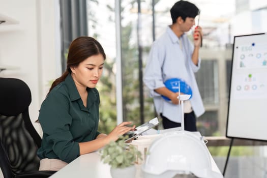 A female engineer attentively works on her tablet, analyzing project details while her colleague engages in the background, showcasing a collaborative workspace.