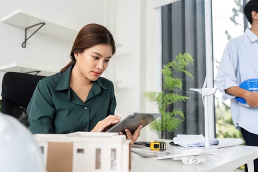 A woman in a green shirt focuses on her tablet, analyzing data for a sustainable architecture project in a modern office.