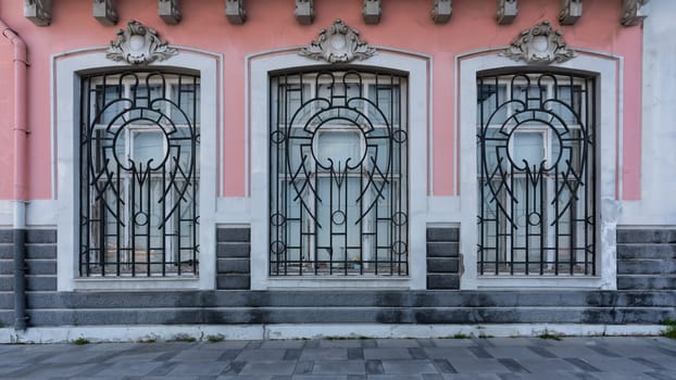 Facade of a pink building with three windows featuring intricate geometric and curved metal grilles. White trim, grey stone steps, sidewalk