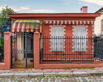 Exterior view of a small orange house with white shutters, black metal fence, and striped awnings on a cobblestone street, at daytime