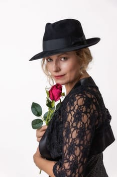 Middle aged woman in a black lace dress and hat holding a red rose in her hand is posing on a white background.