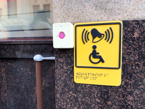 Bright yellow accessibility button for disabled assistance with Braille text and signal icon mounted on granite wall near entrance, promoting inclusive urban infrastructure
