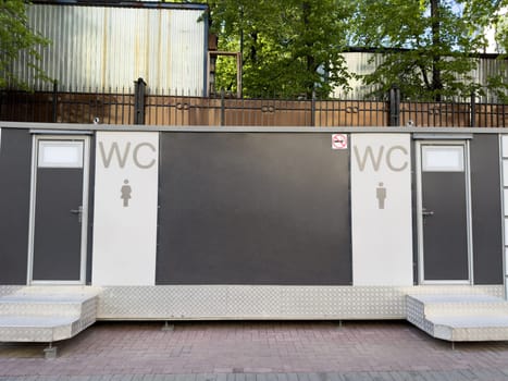 Modern outdoor public restroom unit with separate entrances for men and women, equipped with metal steps, clear WC signage and no smoking symbol located in urban park setting