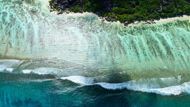 Waves reach the clear sandy shoreline bordered by lush green vegetation from an aerial perspective. Seychelles, La Digue.