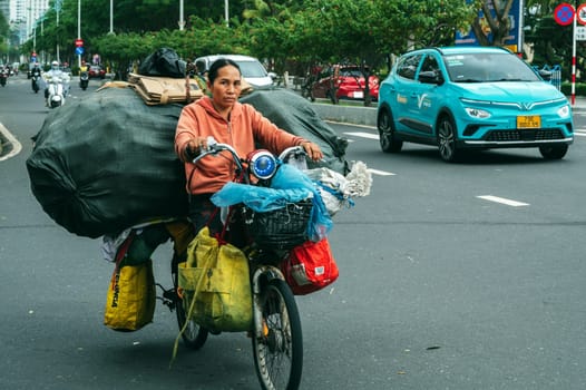An elderly Vietnamese woman in traditional closed clothes and a hat carries garbage on an old bicycle along a city street. Nha Trang, Vietnam - December 14, 2024