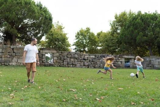 Two girls playing soccer in a park while their father watches them