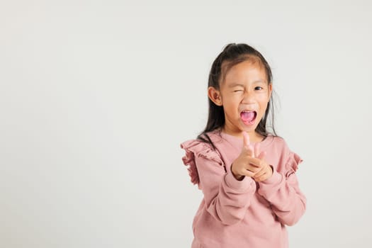 Cheerful Asian young kid girl excitedly makes two gun finger gesture to the camera with a friendly and cheeky expression. Isolated on white background, kindergarten child exudes positivity and joy