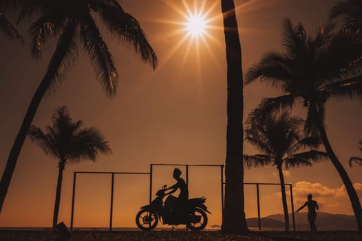 Silhouette of a Vietnamese man on a motorcycle on the embankment with palm trees by sea at sunrise on summer morning in Vietnam