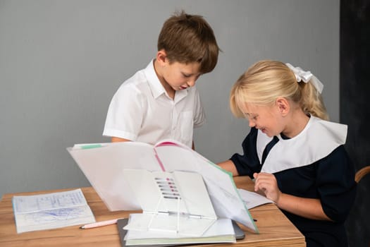 A boy and a girl in school uniforms are sitting at a table, looking at books and writing in their notebooks.