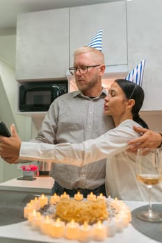 Here's a caption that fits the criteria: A couple, wearing party hats, embraces while taking a selfie in their kitchen. A birthday cake with lit candles sits on the counter.