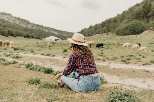 A woman in a plaid shirt and jeans sits in a field with a hat on. She is looking at a herd of sheep