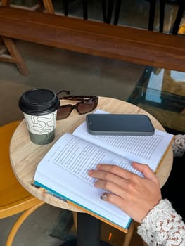 A person relaxes with a book at a cafe table, enjoying coffee and surrounded by sunglasses and a smartphone.