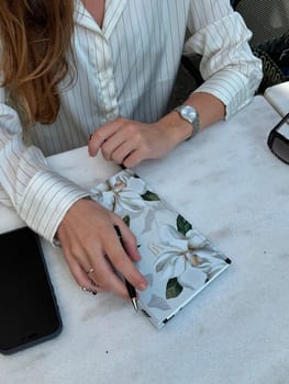 A woman sits at a cafe table, writing in a floral notebook while casually holding a pen. Afternoon light illuminates her workspace.