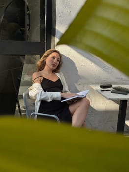 A woman sits relaxed at a table, reading a book while basking in the sunlight surrounded by green leaves.