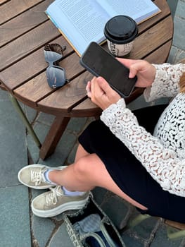 A person relaxes at a wooden table, sipping coffee and looking at their phone while reading a book in the afternoon.