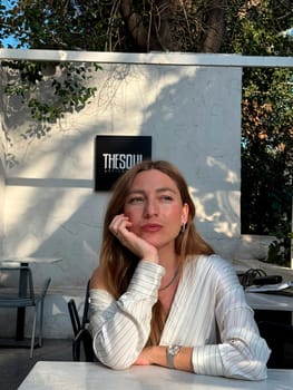 A woman sits at a table in an elegant cafe, resting her chin on her hand while enjoying a tranquil afternoon outdoors.