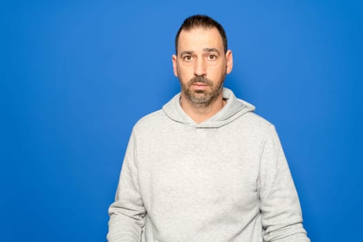 Hispanic man with a beard in his 40s wearing a gray sweatshirt looks straight ahead with a serious and worried expression, isolated on a blue studio background