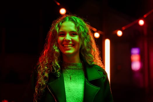 A young woman with curly hair is smiling brightly, surrounded by vibrant neon lights in an urban area at night. The atmosphere is lively and festive, showcasing outdoor decorations.