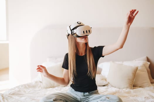 A woman with long hair enjoys a virtual reality experience in her bedroom. Sitting cross-legged on her bed, she uses a headset and reaches out as if interacting with a digital environment.