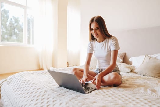 A young woman sits on a bed in a sunlit room, engaging with her laptop. She appears focused and relaxed, surrounded by soft textiles and natural light, creating a cozy atmosphere.