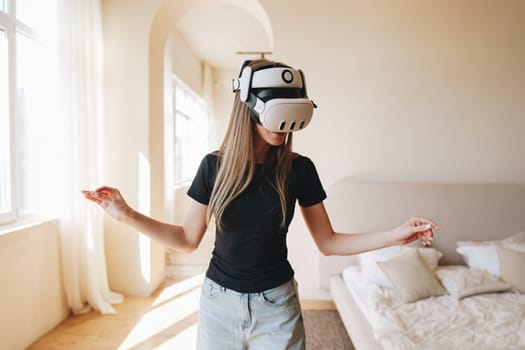 A young woman immersed in virtual reality gaming moves her hands gracefully while wearing a VR headset in a cozy, sunlit room. The background features light-colored furniture.
