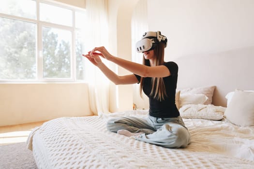 A young woman wearing a virtual reality headset engages with an immersive experience in her brightly lit bedroom. She sits cross-legged on a soft bed, captivated by the virtual world.