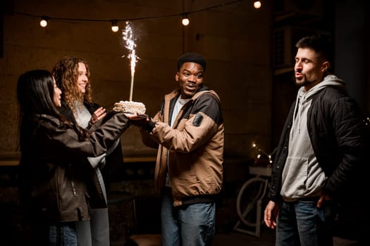 Friends celebrate together at an evening gathering, one person holding a cake adorned with a sparkling candle. Laughter and smiles fill the atmosphere, creating a lively moment.