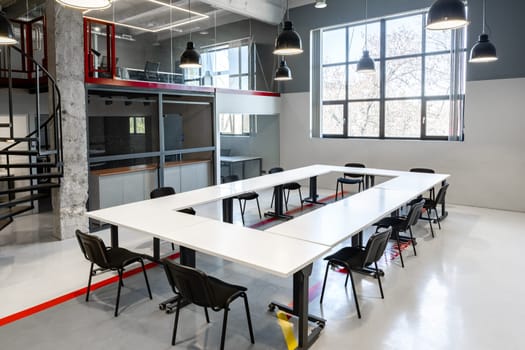 A spacious coworking area features a large white table surrounded by chairs. Large windows let in natural light, enhancing the modern design and atmosphere suitable for teamwork.