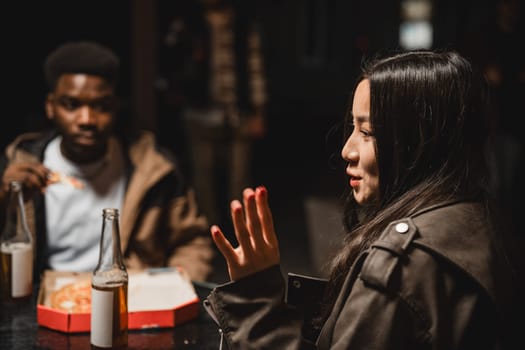 Two friends share a meal at a dimly-lit table, engaging in lively conversation while enjoying drinks and takeaway food in an urban environment.