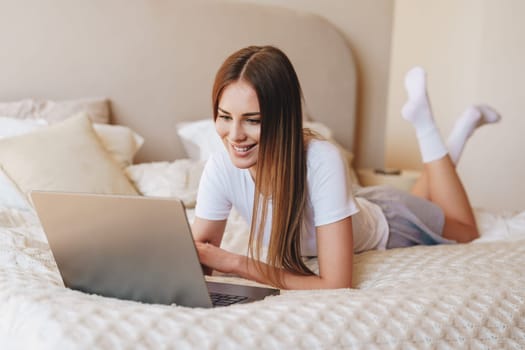 A young woman relaxes on her bed, smiling as she uses a laptop. Soft light fills the cozy room, creating a comfortable atmosphere for her online activities.