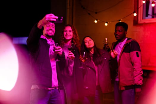 A group of four friends with drinks pose for a selfie in a cozy outdoor setting illuminated by string lights. Their joyful expressions capture the fun atmosphere of their gathering.