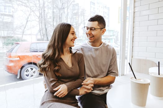 A couple sits closely together at a cafe, laughing and sharing a happy moment. They enjoy their drinks while sunlight filters through the windows, creating a warm atmosphere.