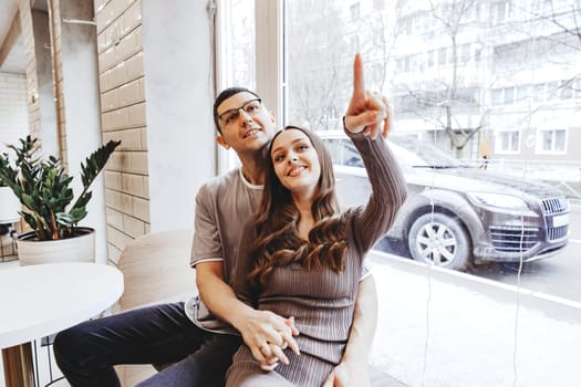 A couple sits closely in a cafe, with one partner pointing excitedly toward the snowy view outside. The atmosphere is warm and inviting, perfect for a winter day.