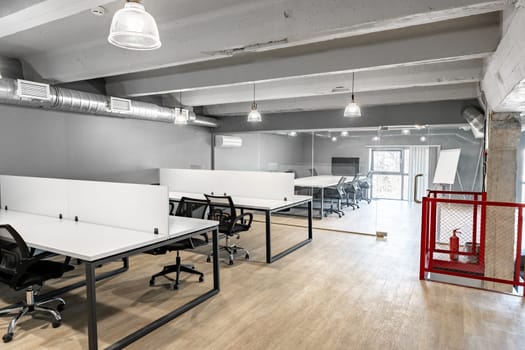 An open coworking area features several desks with ergonomic chairs in a spacious, well-lit setting. Large windows provide natural light, enhancing productivity.