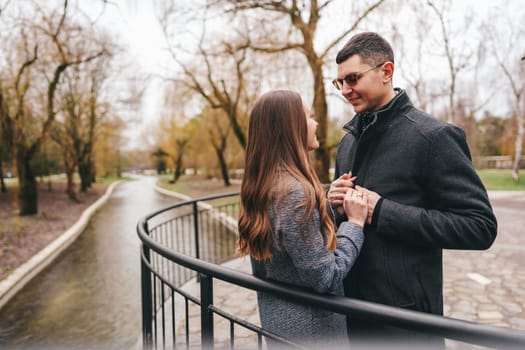 A couple stands close together by a winding river in a park, engaging in a tender conversation while surrounded by bare trees. It is a cloudy day in early spring.